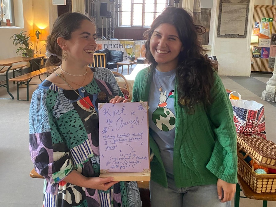 Two smiling women stand inside The Church café-bar in Ipswich, holding a hand-drawn poster for the Knit in the Church event, with knitting baskets and bags of yarn visible nearby